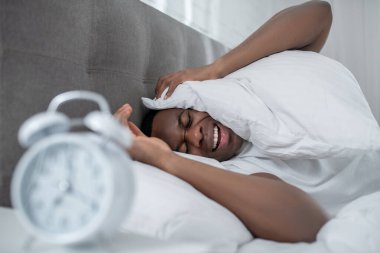 Beyaz tişörtlü genç bir adam yatakta yatıyor ve çalar saatin sesinden rahatsız oluyor.