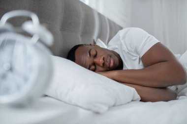 Beyaz tişörtlü genç bir adam yatakta yatıyor ve uykulu görünüyor.