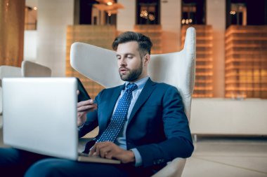 Takım elbiseli bir adam koltukta oturuyor ve dizüstü bilgisayarla çalışıyor.