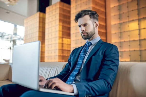 Takım elbiseli sakallı bir adam dizüstü bilgisayarda çalışıyor ve konsantre görünüyor.