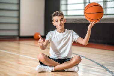 Koyu saçlı bir çocuk yerde oturuyor ve elinde bir top tutuyor.