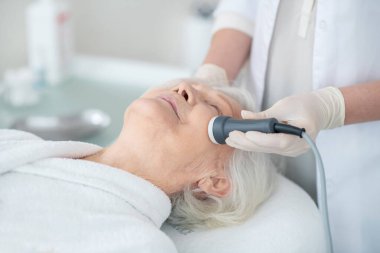 Yaşlı bir müşteriye mikro akım terapisi yapan bir güzellik uzmanı.