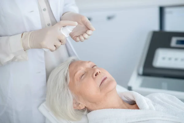 Cosmetologist trabalhando com um cliente maduro e preparando creme facial para aplicação — Fotografia de Stock