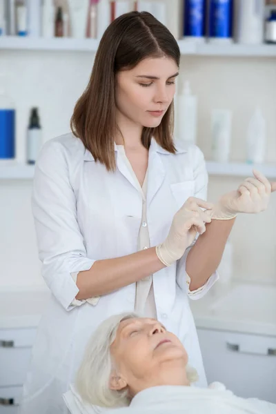Kosmetolog v bílém plášti obléká sterilní rukavice — Stock fotografie