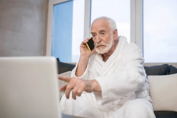 Homem barbudo de cabelos grisalhos com roupão branco falando ao telefone e parecendo ocupado — Fotografia de Stock