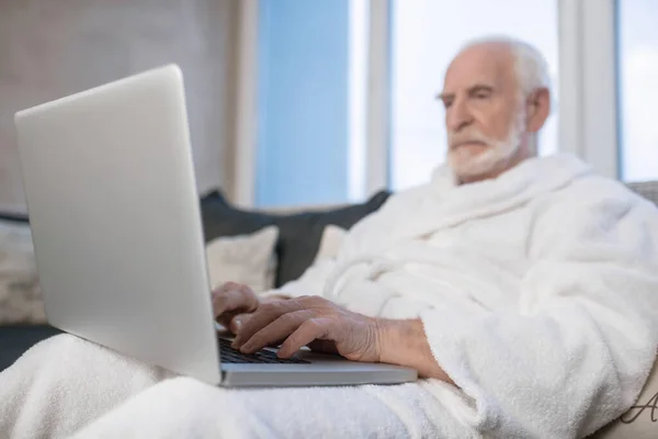 Ősz hajú, szakállas férfi fehér köntösben, aki laptopon dolgozik és komolynak tűnik. — Stock Fotó