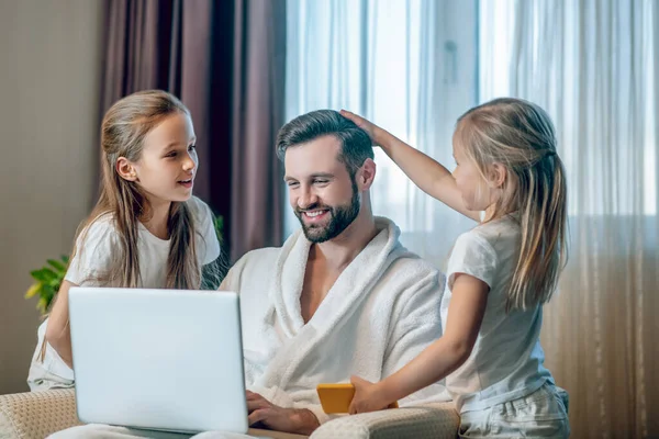 Fiatal apa ül egy laptoppal, és lányai állnak hime mellett. — Stock Fotó