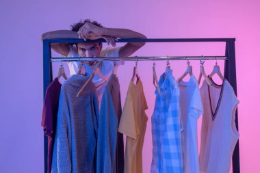 Afro-Amerikalı genç adam elbise askılarının yanında duruyor ve düşünceli görünüyor.