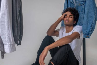 Kıvırcık saçlı genç adam askıların yanında oturuyor ve üzgün görünüyor.