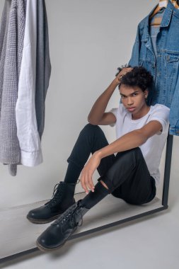 Kıvırcık saçlı genç adam askıların yanında oturuyor ve üzgün görünüyor.