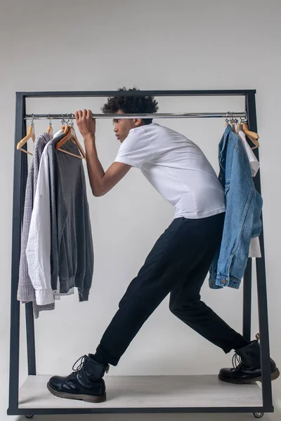 Africano americano jovem cara segurando cabides com roupas e inclinando-se para a frente — Fotografia de Stock