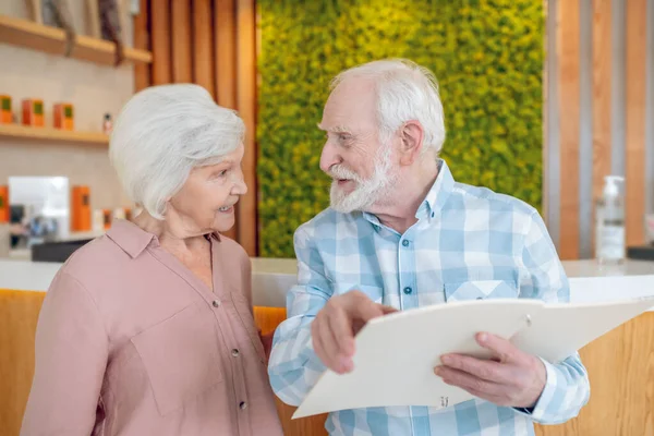 Gray-haired couple looking trhough the price list in spa center and looking excited — Stock Photo, Image