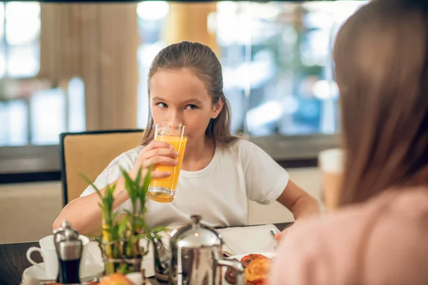 Tini lány ül az asztalnál és narancslevet iszik — Stock Fotó
