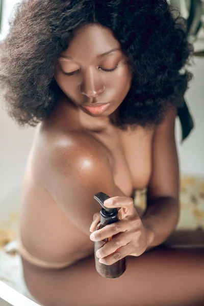 Naked dark-skinned young woman applying body lotion on her body — Stock Photo, Image