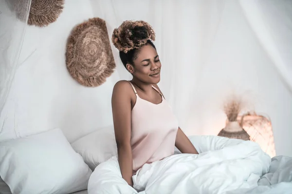 Joyful dark-skinned woman in nightie sitting on bed — Stock Photo, Image