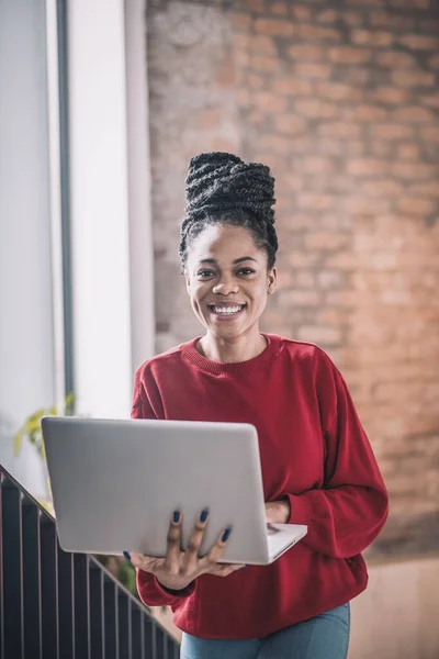 Kırmızı tişörtlü, laptoplu, gülümseyen ve ofiste yürüyen siyah bir kadın. — Stok fotoğraf