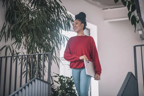 Mujer negra en camisa roja con un portátil sonriendo y caminando en la oficina — Foto de Stock