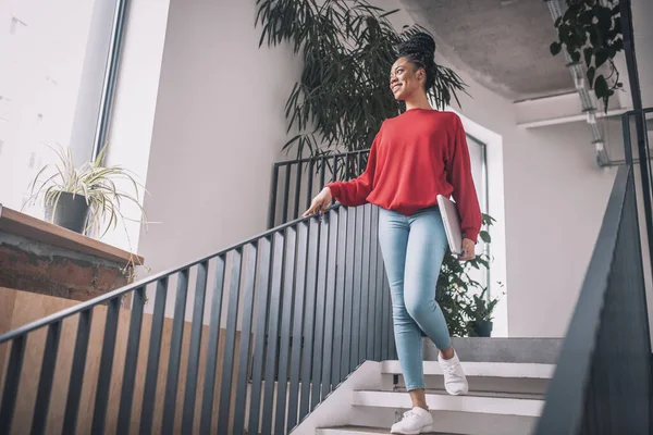 Mujer negra en camisa roja con un portátil sonriendo y caminando en la oficina — Foto de Stock