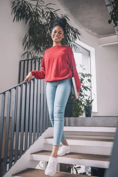 Mujer negra en camisa roja con un portátil sonriendo y caminando en la oficina —  Fotos de Stock