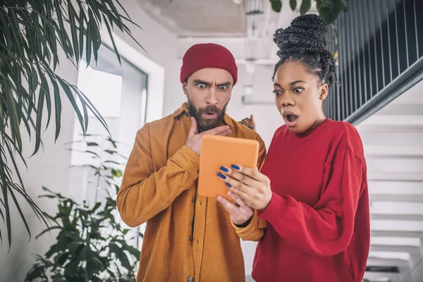 Un hombre con un sombrero rojo y una mujer negra mirando sorprendido whule viendo algo en una tableta —  Fotos de Stock