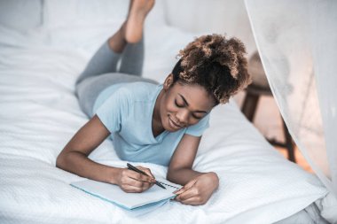 Yatağa uzanmış, deftere yazı yazan bir kadın.