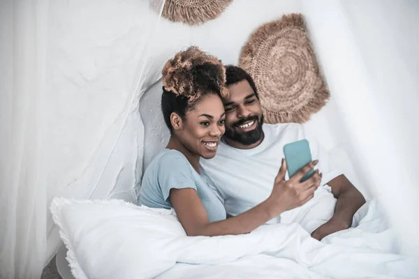 Žena s tmavou pletí a muž s selfie v posteli — Stock fotografie