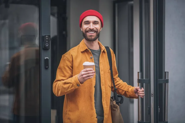 Man i röd hatt och orange jacka med en kaffekopp i händerna ser positivt — Stockfoto