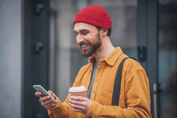 Hombre con un sombrero rojo y chaqueta naranja con una taza de café en las manos mirando alegre —  Fotos de Stock