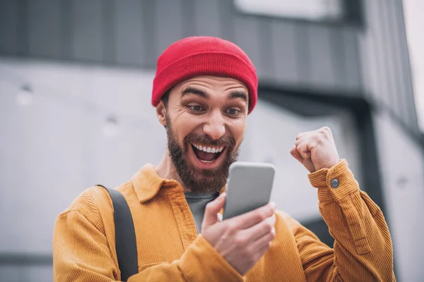 Joven barbudo con un sombrero rojo mirando su teléfono y sintiéndose feliz — Foto de Stock