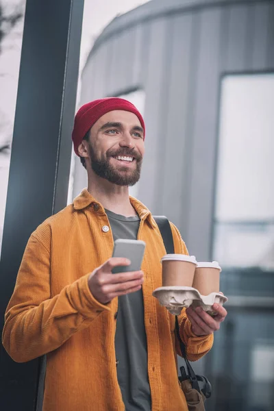 Joven barbudo con un sombrero rojo sosteniendo tazas de café y luciendo alegre —  Fotos de Stock