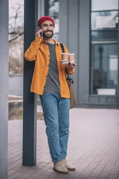 Joven barbudo con un sombrero rojo hablando por teléfono y llevando las tazas de café —  Fotos de Stock