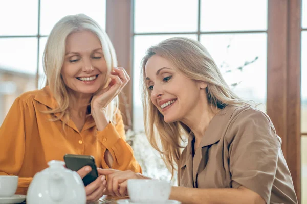 Dos señoras lindas pasar tiempo juntos y discutir fotos en un teléfono inteligente —  Fotos de Stock