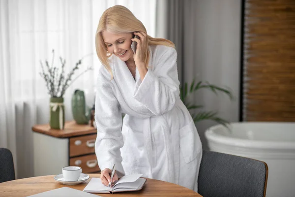 Långhårig kvinna i en vit badrock pratar i telefon och antecknar — Stockfoto