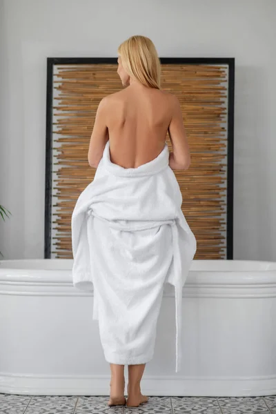 Egy gyönyörű nő fehér fürdőköpenyben, aki meztelenül mutatja a hátát. — Stock Fotó