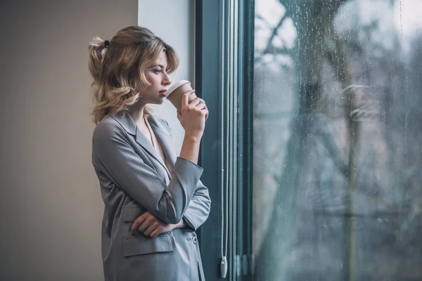 Mujer pensativa bebiendo café de pie cerca de la ventana —  Fotos de Stock