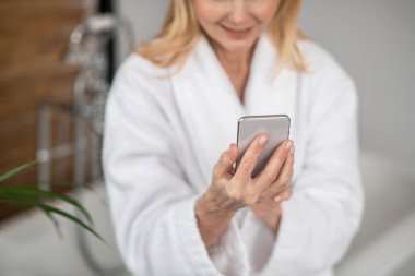 Elinde akıllı bir telefonla beyaz banyo bornozu giymiş bir kadın.
