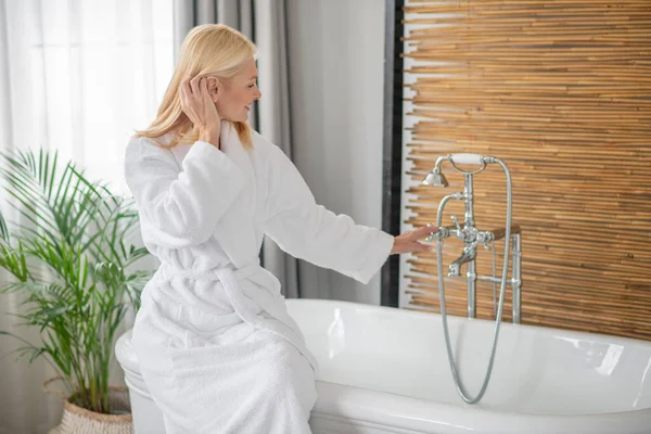 Blonde hübsche Frau in weißem Bademantel bereitet die Badewanne vor — Stockfoto