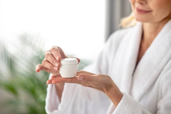 Una mujer con una bata blanca sosteniendo un frasco de crema facial — Foto de Stock