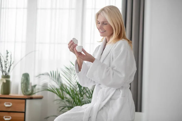 Eine blonde Frau im weißen Bademantel hält ein Glas mit einer neuen Creme in der Hand — Stockfoto