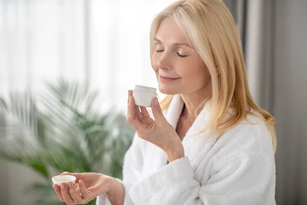 Una mujer rubia con una bata blanca sosteniendo un frasco de una nueva crema — Foto de Stock