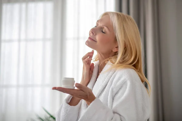 Una donna con un accappatoio bianco sembra contenta mentre applica la crema sul viso — Foto Stock