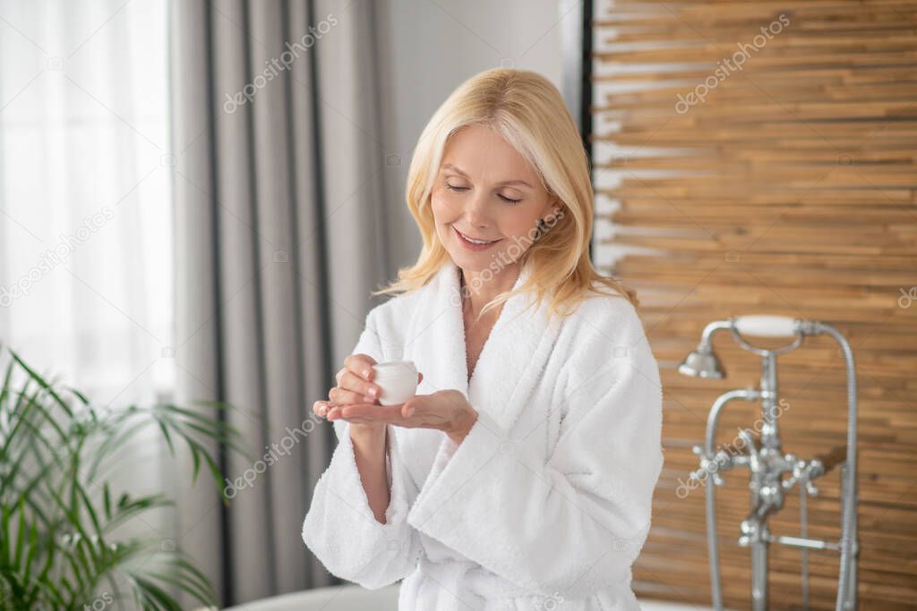 A woman in a white bath robe holding a jar of a facial cream