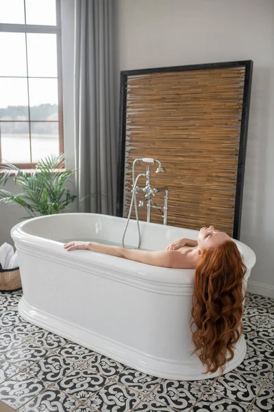 Beautiful ginger woman lying in a bath and looking enjoyed — Stock fotografie