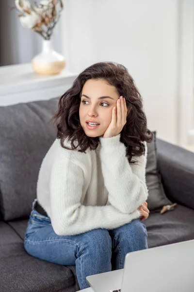 Uma jovem mulher em uma camisola branca sentada no sofá e olhando para o lado — Fotografia de Stock