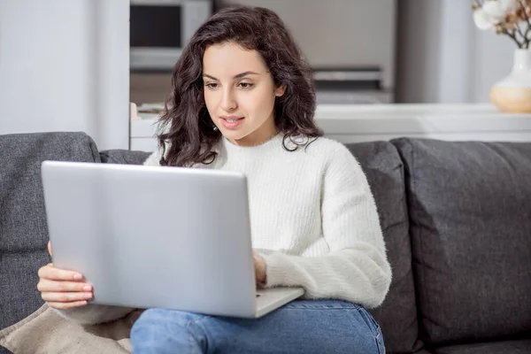 Uma jovem mulher em uma camisola branca sentada no sofá com um laptop — Fotografia de Stock