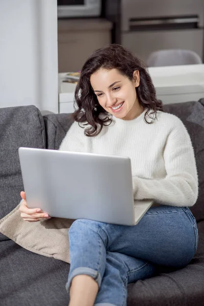 Una giovane donna con un maglione bianco seduta sul divano con un computer portatile — Foto Stock