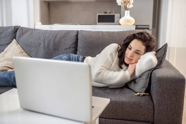 Uma mulher de camisola branca deitada no sofá e assistindo algo online — Fotografia de Stock