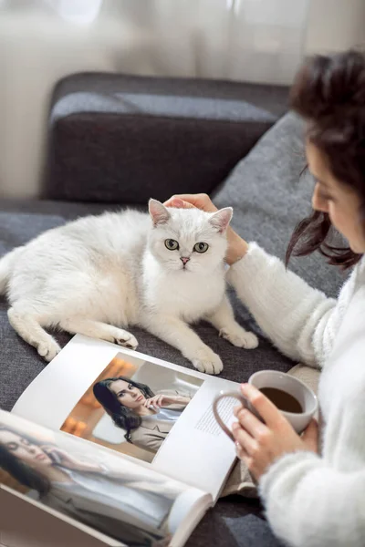 Eine Frau ruht sich zu Hause aus, liest Zeitschriften und streichelt ihre Katze — Stockfoto