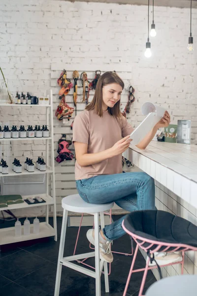 Una chica en una camiseta beige sentada en una tienda de mascotas y mirando a través de la lista de precios —  Fotos de Stock