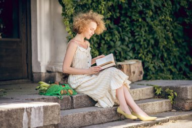 Genç bir kadın elinde kitapla merdivenlerde oturuyor.